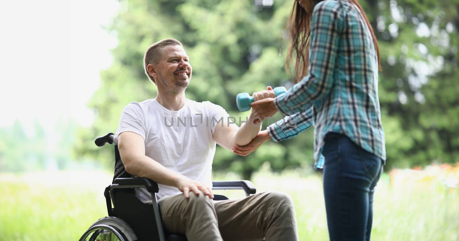 Female physiotherapist helps young man in wheelchair to lift weight of arms in park by kuprevich
