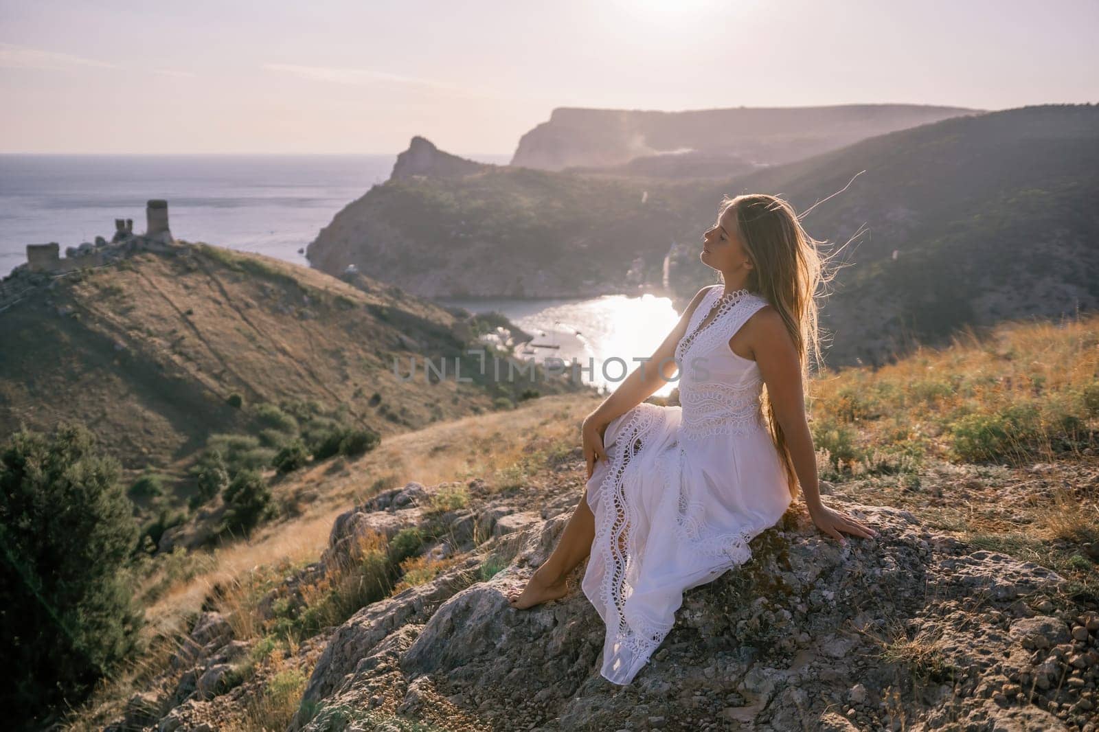 A woman in a white dress is sitting on a rock overlooking a body of water. She is enjoying the view and taking in the scenery
