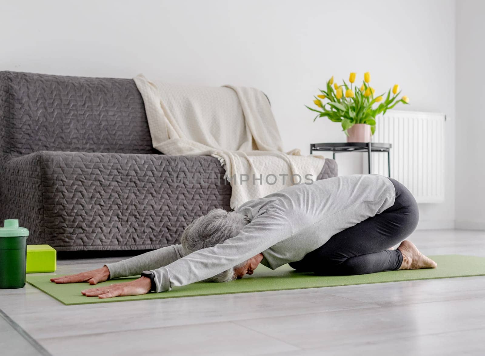 Grey-Haired Woman In baby Pose Practices Yoga At Home On The Floor In A Bright Living Room