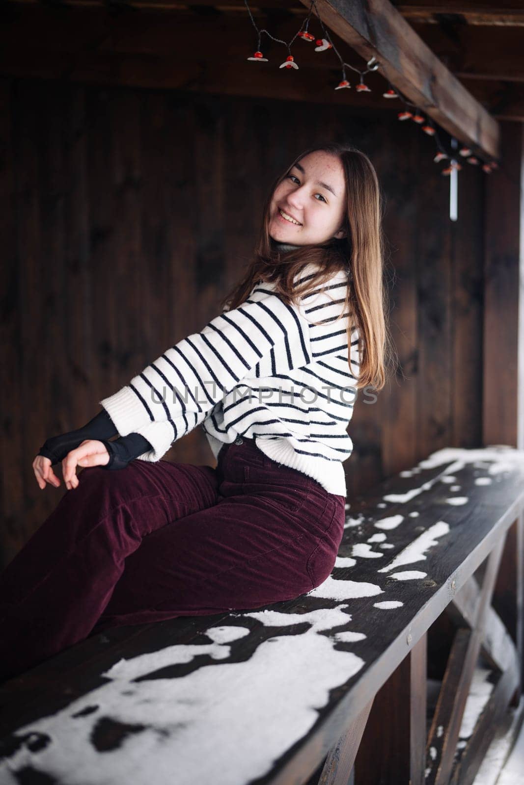 Schoolgirl wearing striped sweater and corduroy trousers smiling while sitting on a snowy bench during winter
