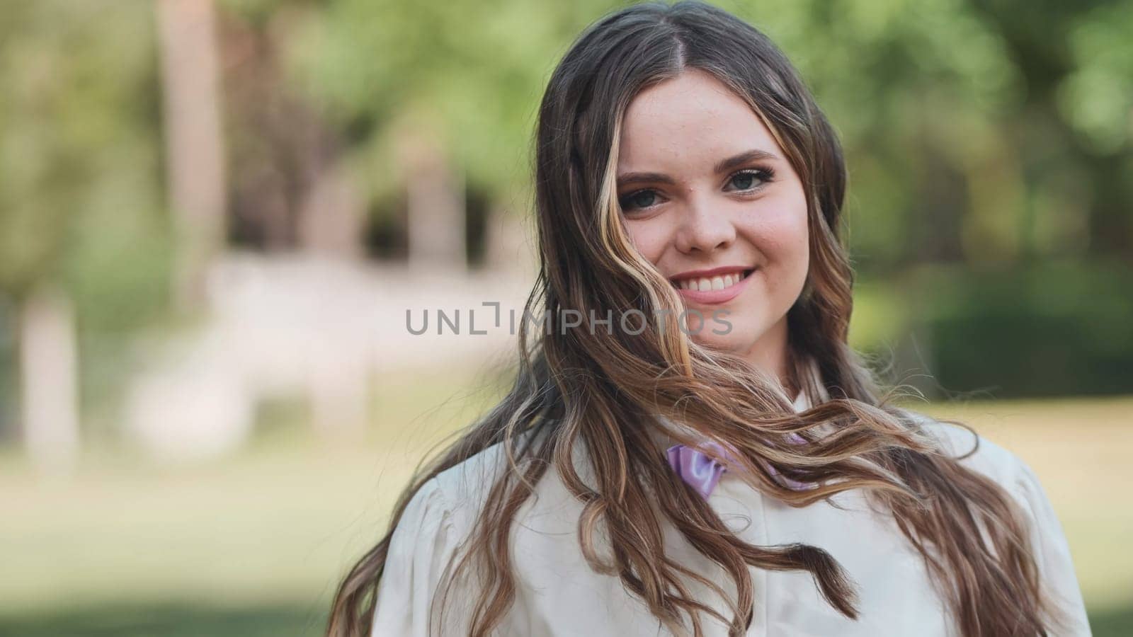 Portrait of a cheerful young woman with long, flowing hair, enjoying a sunny day in a park, wearing a stylish white shirt