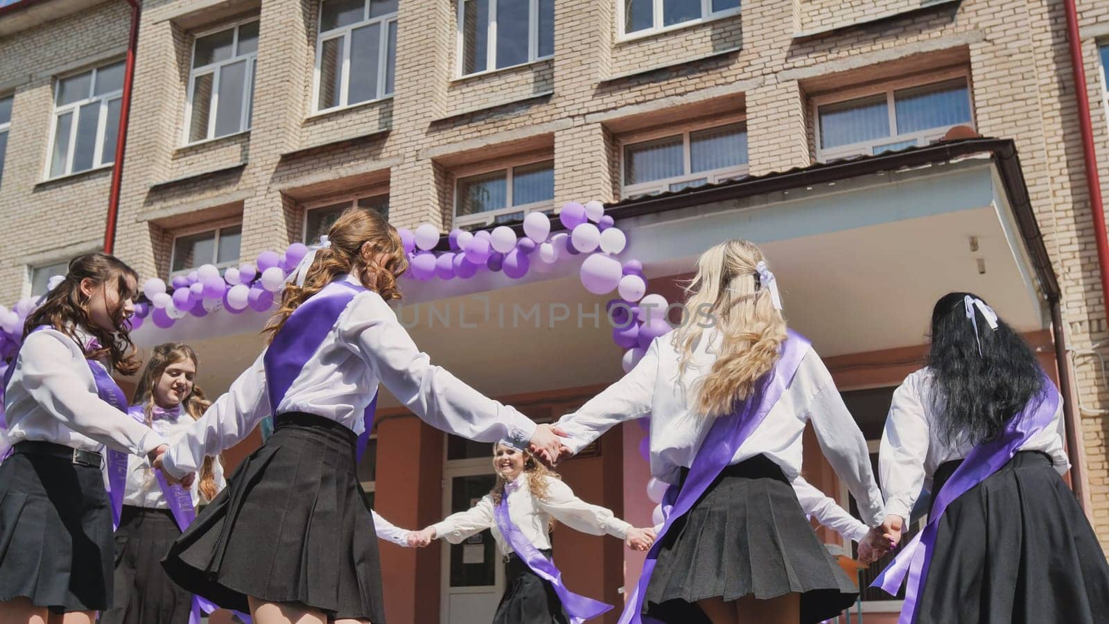 Group of happy female students wearing purple ribbons dancing in a circle in front of their school on their last school day
