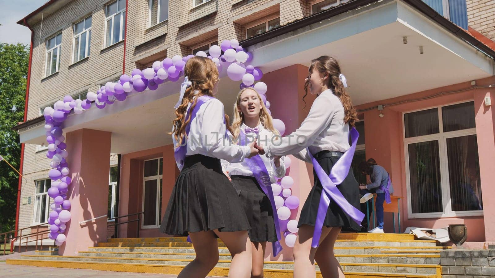 Three cheerful female students with purple ribbons are dancing and celebrating their last school day in front of the school building decorated with balloons.