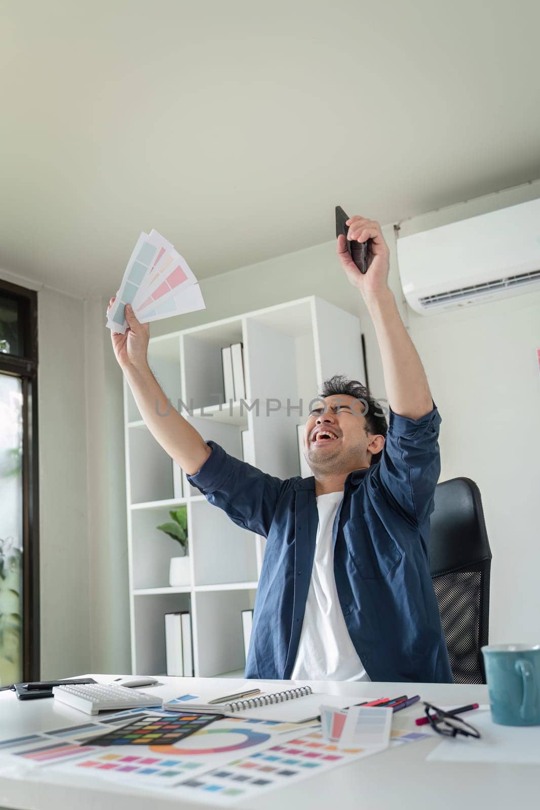 A graphic designer joyfully celebrates a successful project in a modern office, holding color swatches and a smartphone.