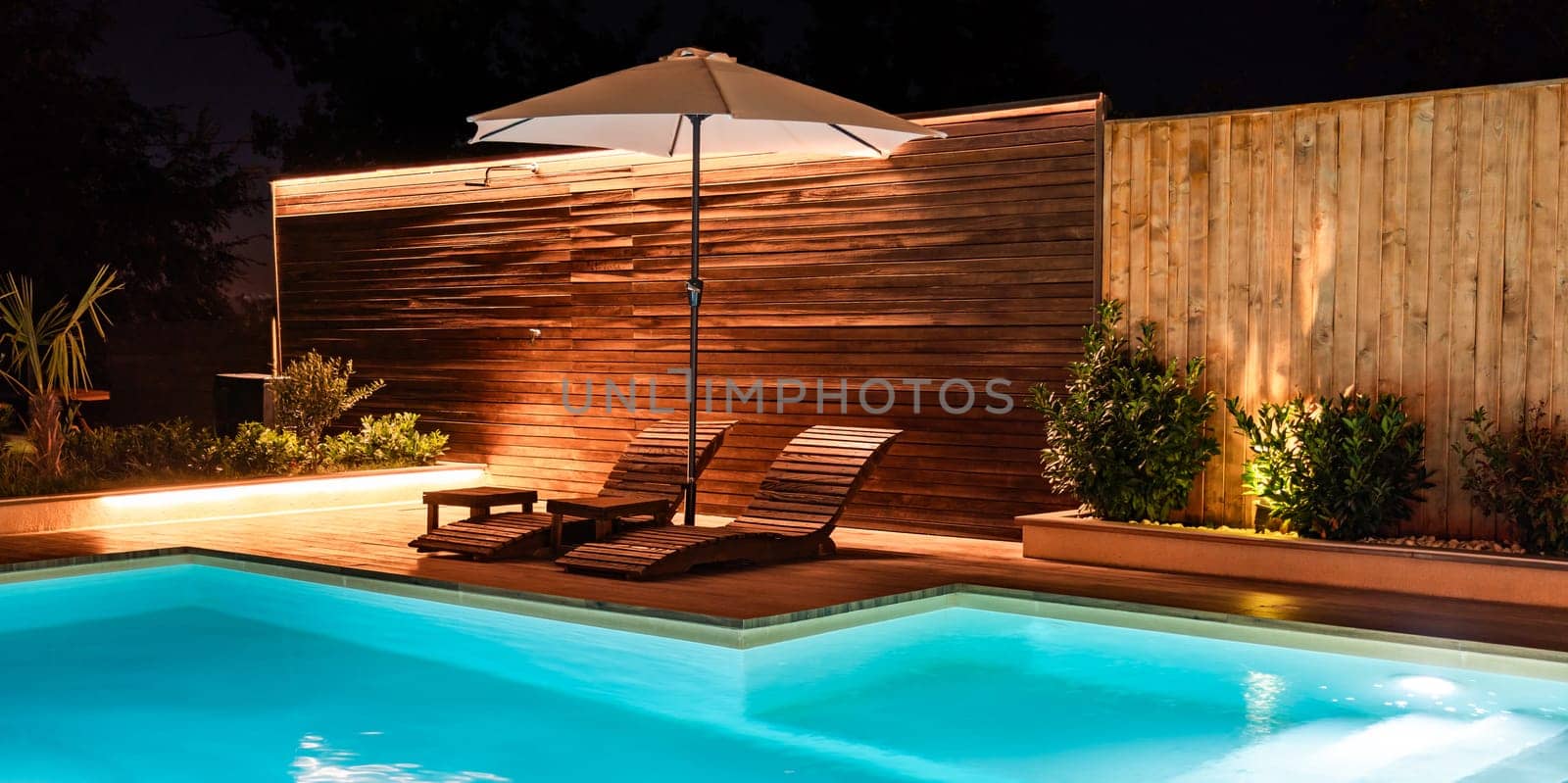 Modern illuminated swimming pool with wooden lounge chairs and a cozy umbrella creating a peaceful backyard ambiance. Concept of outdoor comfort and luxury at night