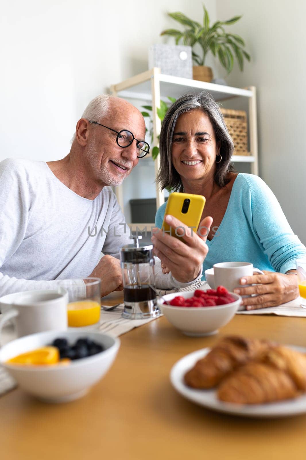 Vertical portrait of happy mature caucasian married couple enjoying healthy breakfast at home looking at phone. Lifestyle concept.