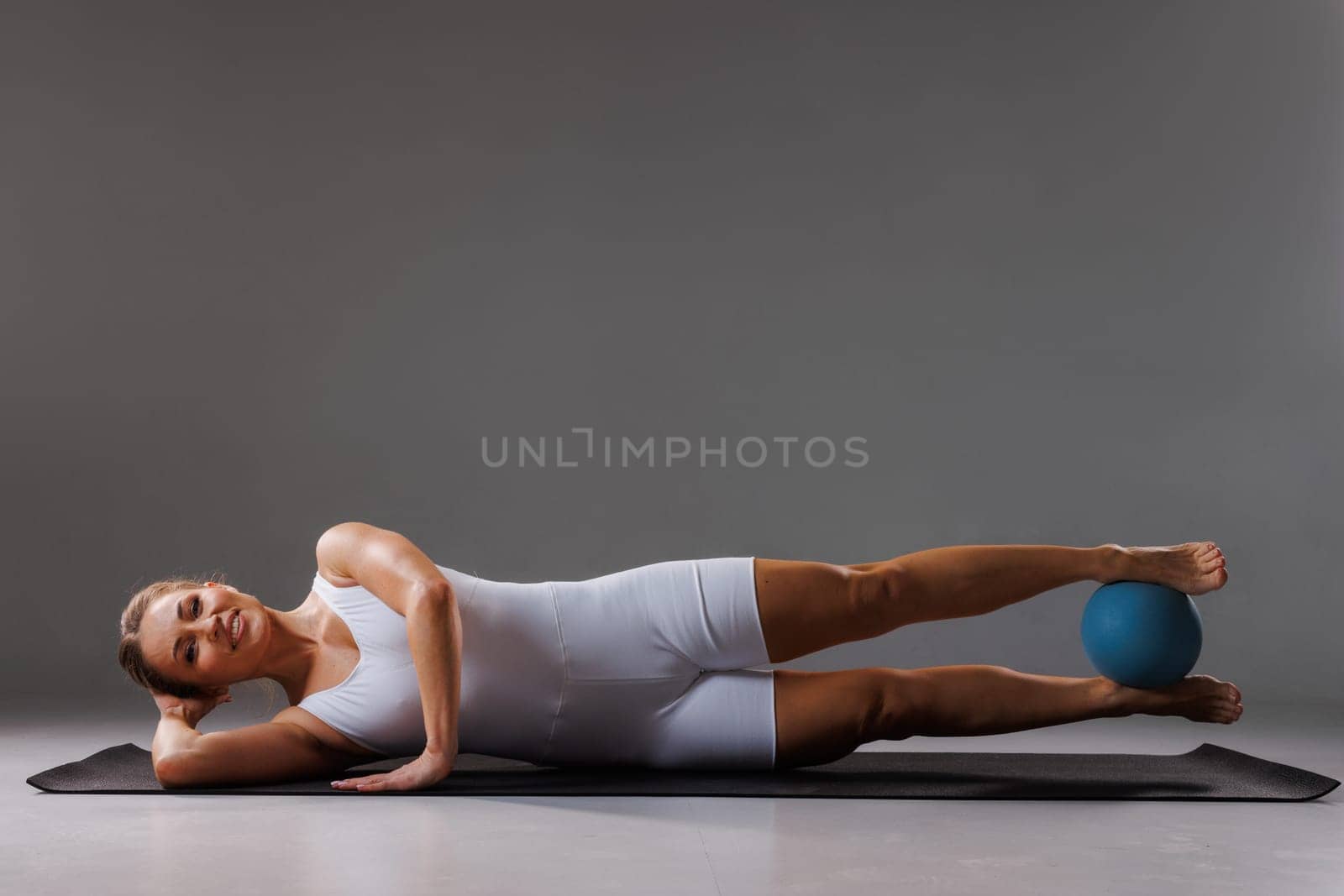 Young woman in white sportswear practicing yoga and stretching with small ball in studio. Athletic woman sitting on mat doing yoga pose.