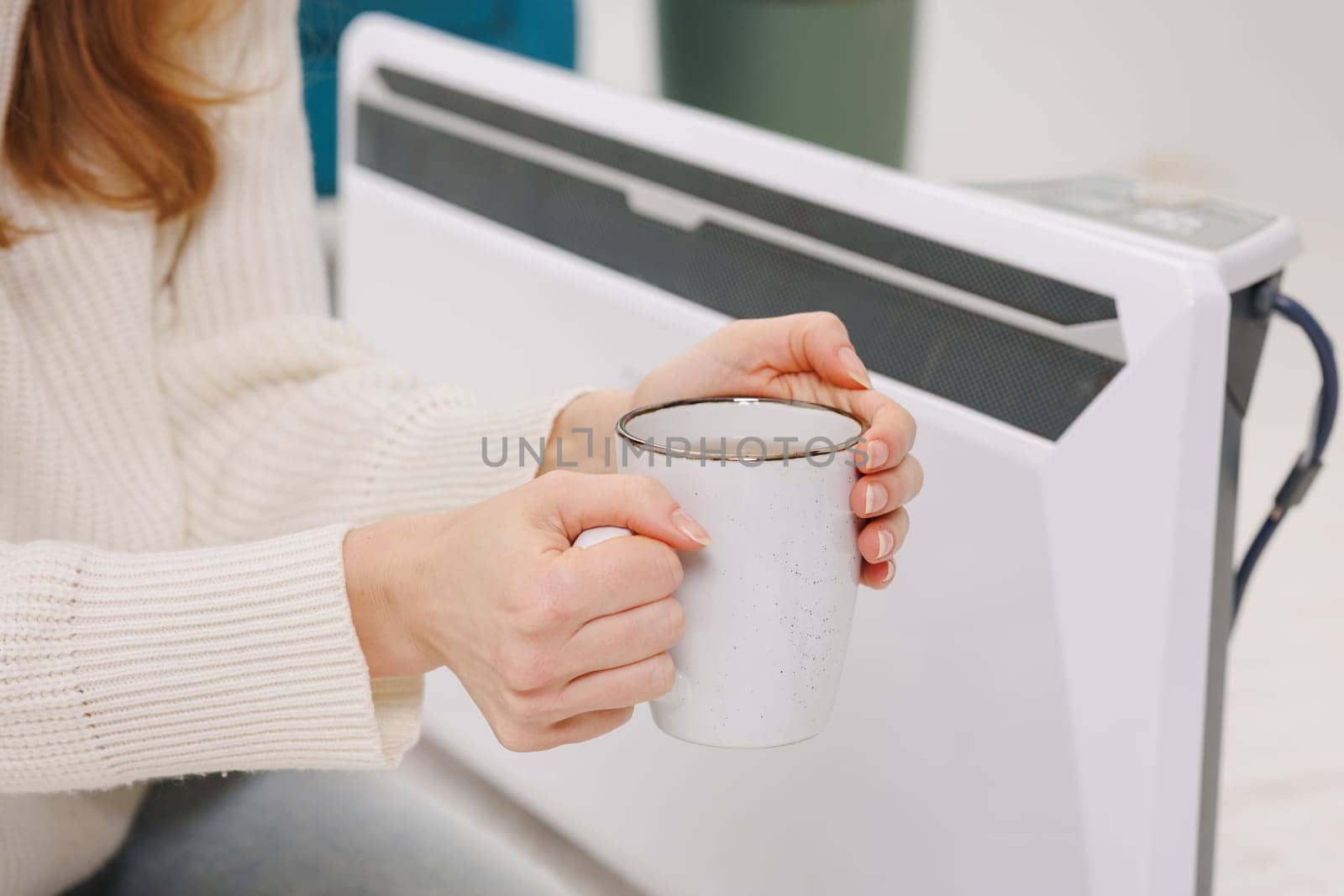 Young woman with warm plaid and tea cup sitting near electric convector heater in bedroom,
