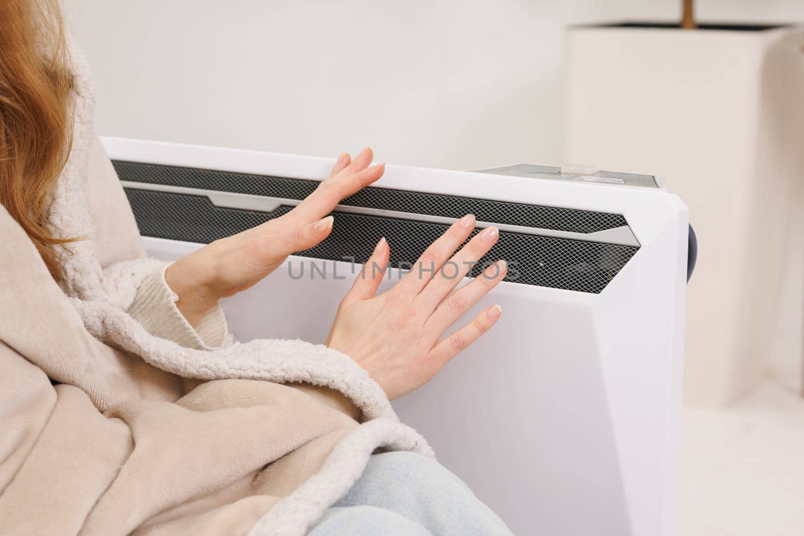 Frozen young woman in mittens warming near radiator at home, closeup.