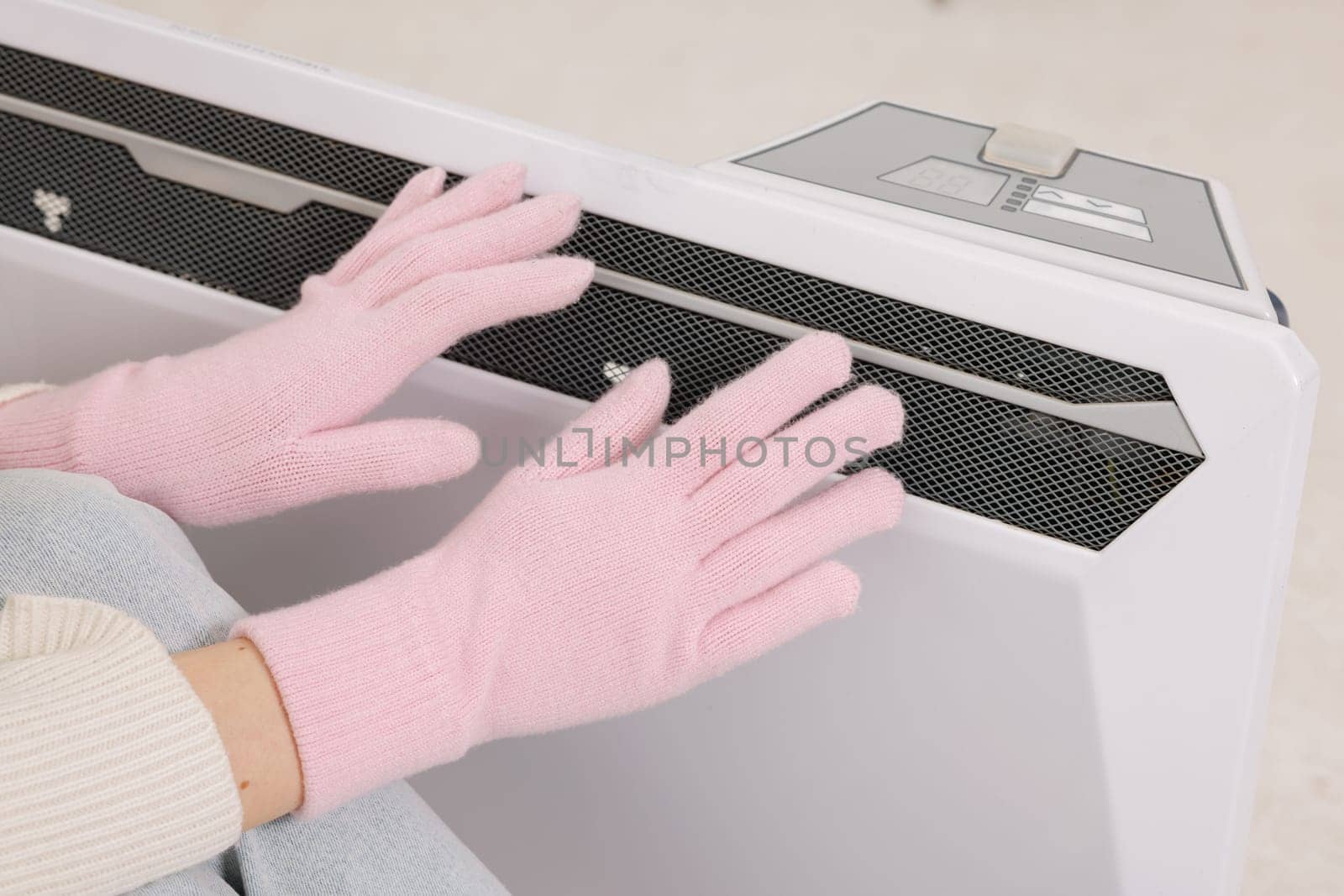 Frozen young woman in mittens warming near radiator at home, closeup.