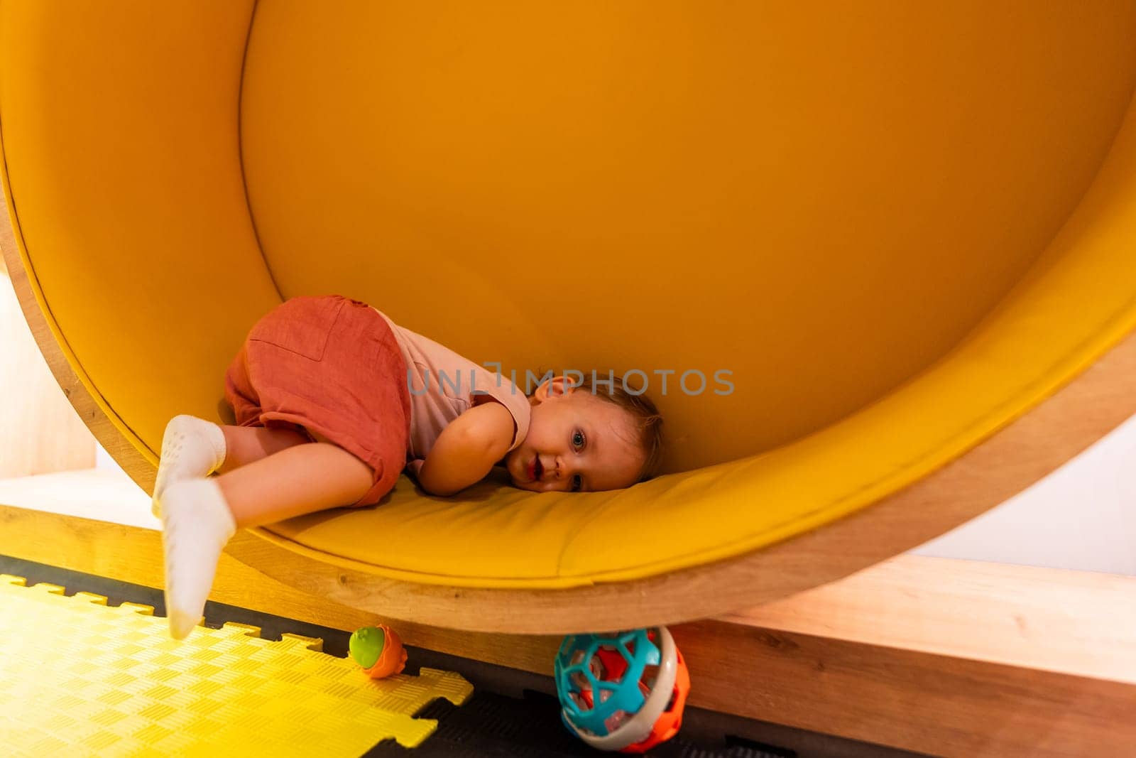 Toddler resting inside a yellow circular play structure in a colorful indoor playroom