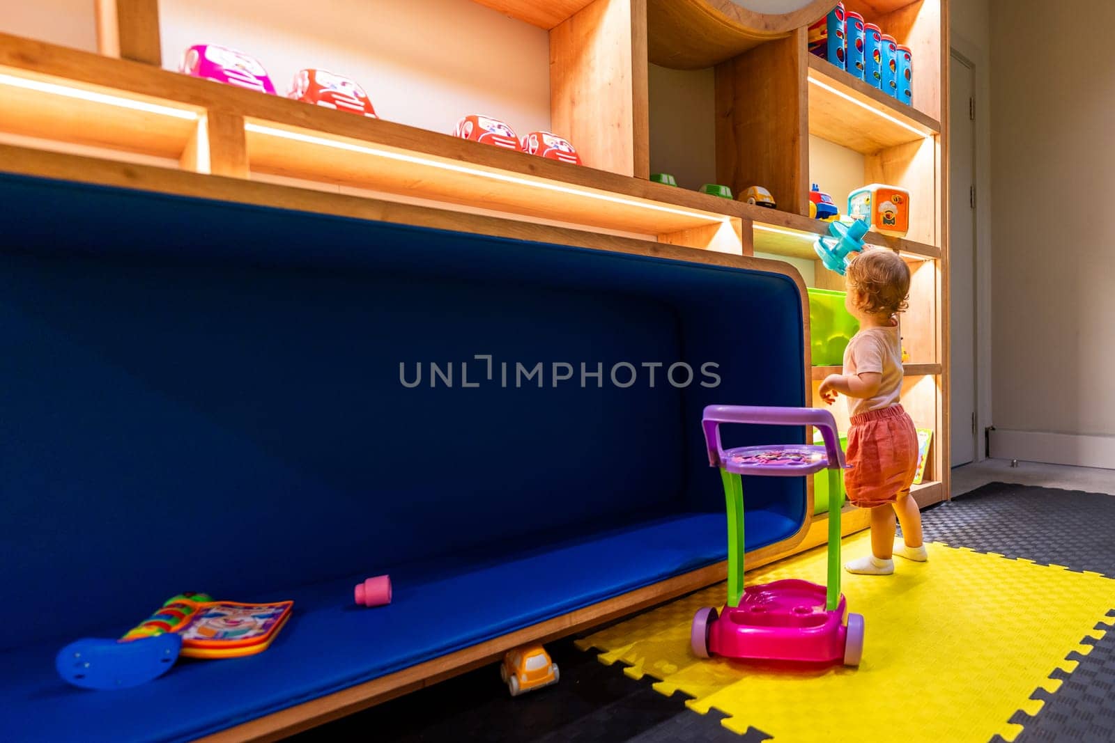 Toddler exploring a colorful playroom with a toy vacuum cleaner and shelves filled with educational toys and games