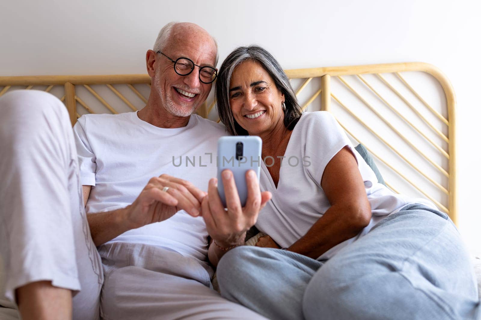 Happy mature caucasian couple looking at mobile phone together relaxing in bed. Lifestyle concept.