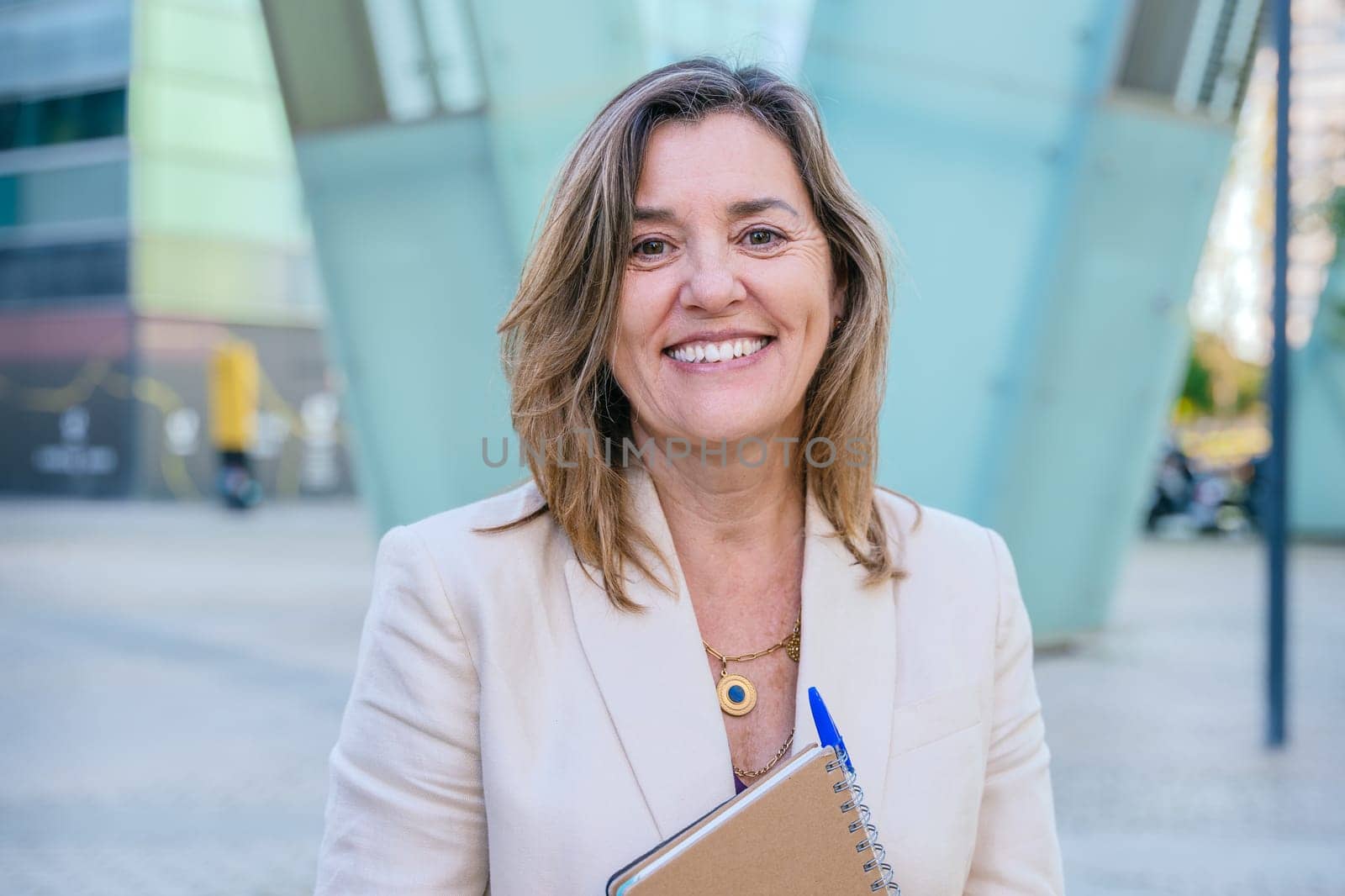 Confident middle-aged businesswoman smiling while holding a notebook. She stands outdoors in an urban setting with modern architecture in the background.