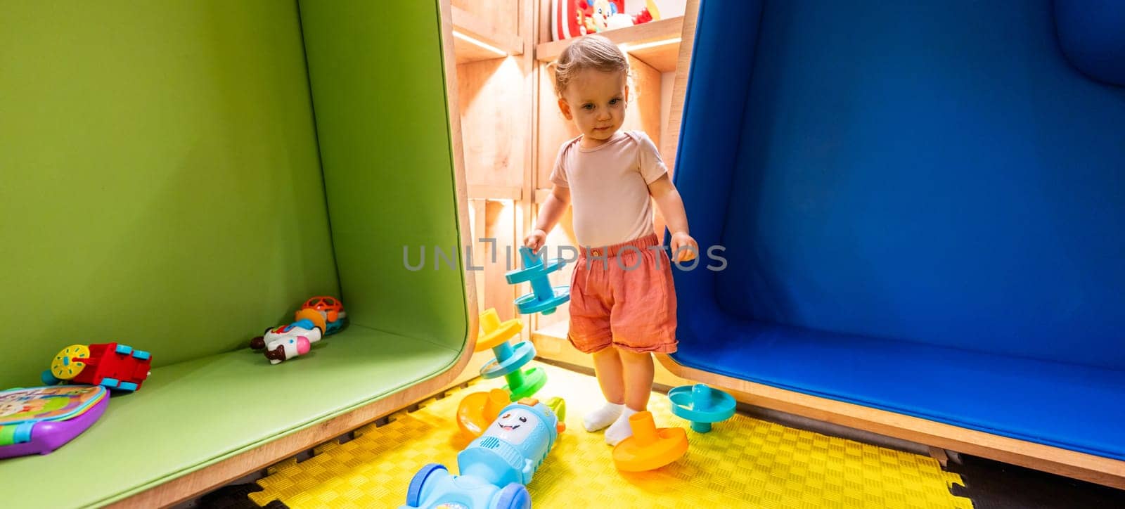 Toddler exploring colorful playroom with educational toys, shelves, and cozy seating areas for creative learning and fun