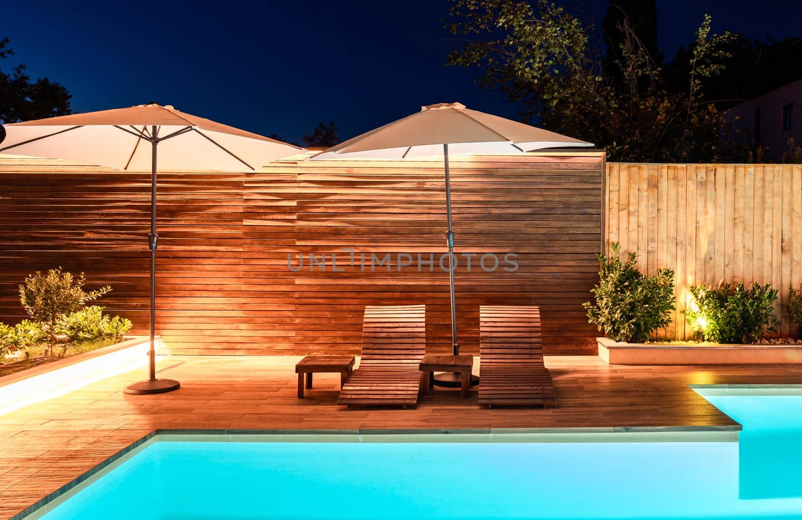Elegant poolside area with lounge chairs and umbrellas at night. Modern outdoor design illuminated with warm lighting