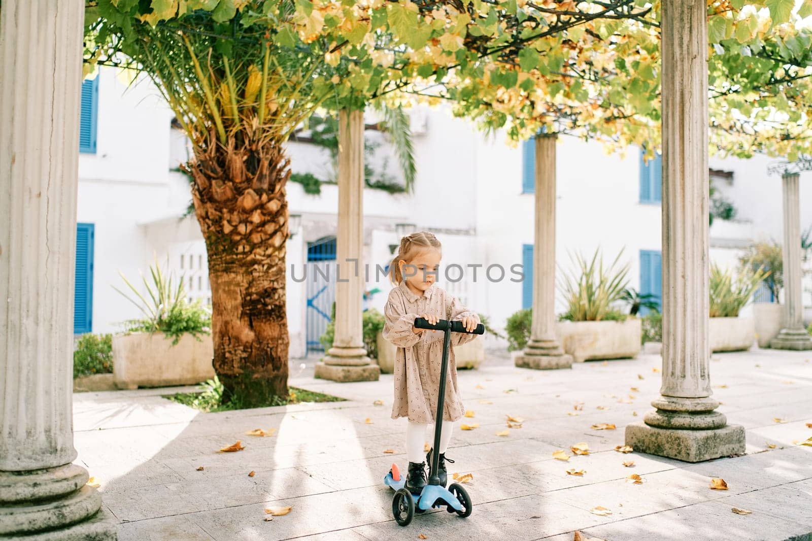 Little girl on a scooter rides between the columns of a pergola in the courtyard of a house. High quality photo