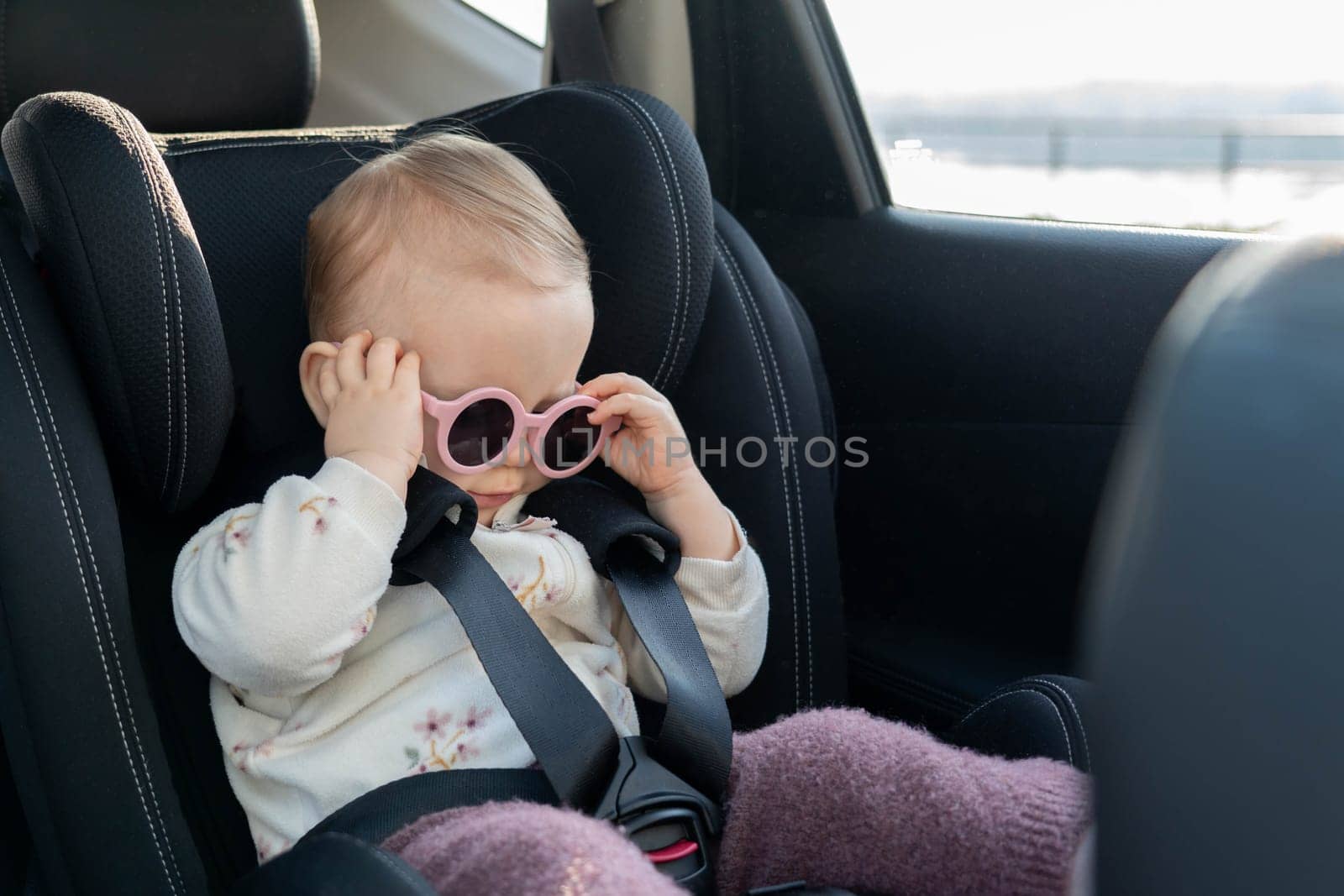 A cute baby wearing pink sunglasses, seated securely in a car seat, looking stylish and ready for adventure. Concept of travel safety, childhood fun, and charming fashion