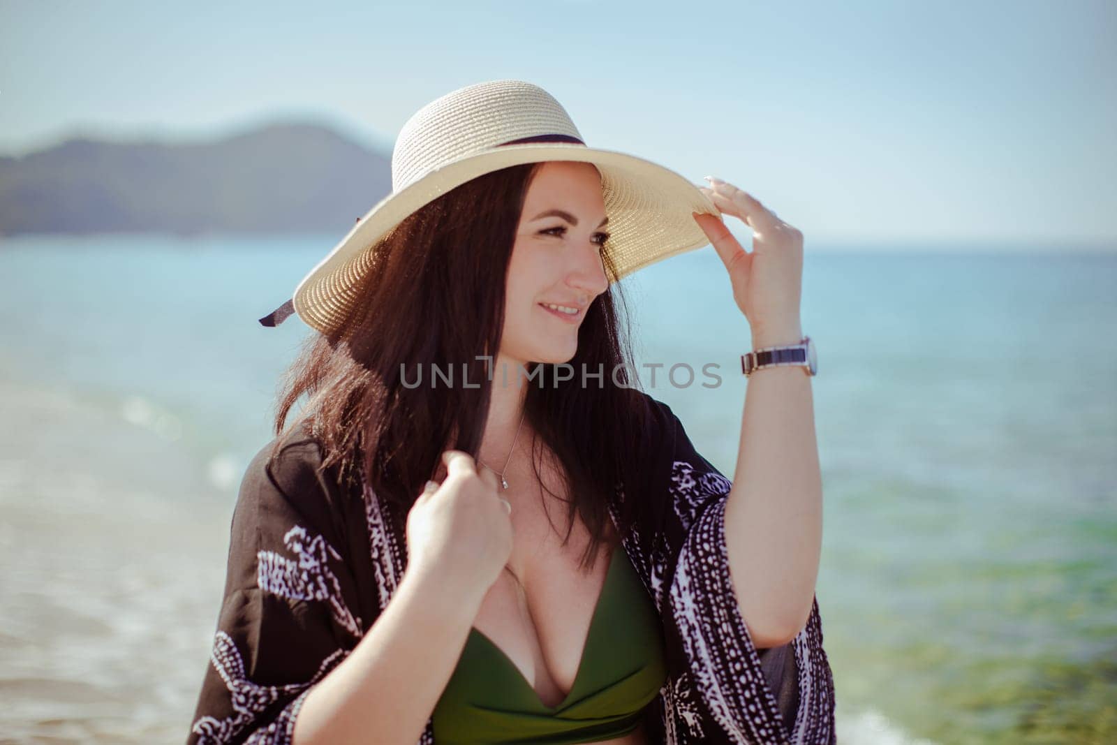 A brunette girl in a beige hat with a large brim stands in a black cape against the background of the sea and mountains