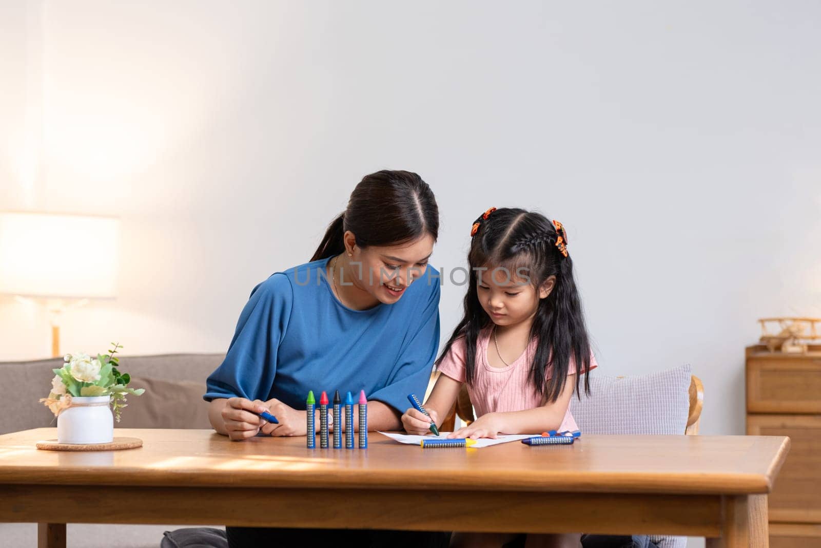 A young family spends their free time together in the living room at home. Mother and little daughter draw pictures with crayons on paper, smiling happy. by wichayada