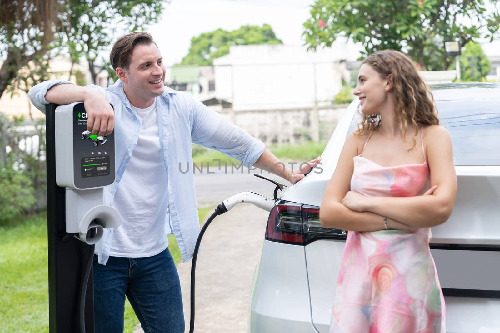 Happy and lovely couple with eco-friendly conscious recharging electric vehicle from EV charging station. EV car technology utilized as alternative transportation for future sustainability. Synchronos