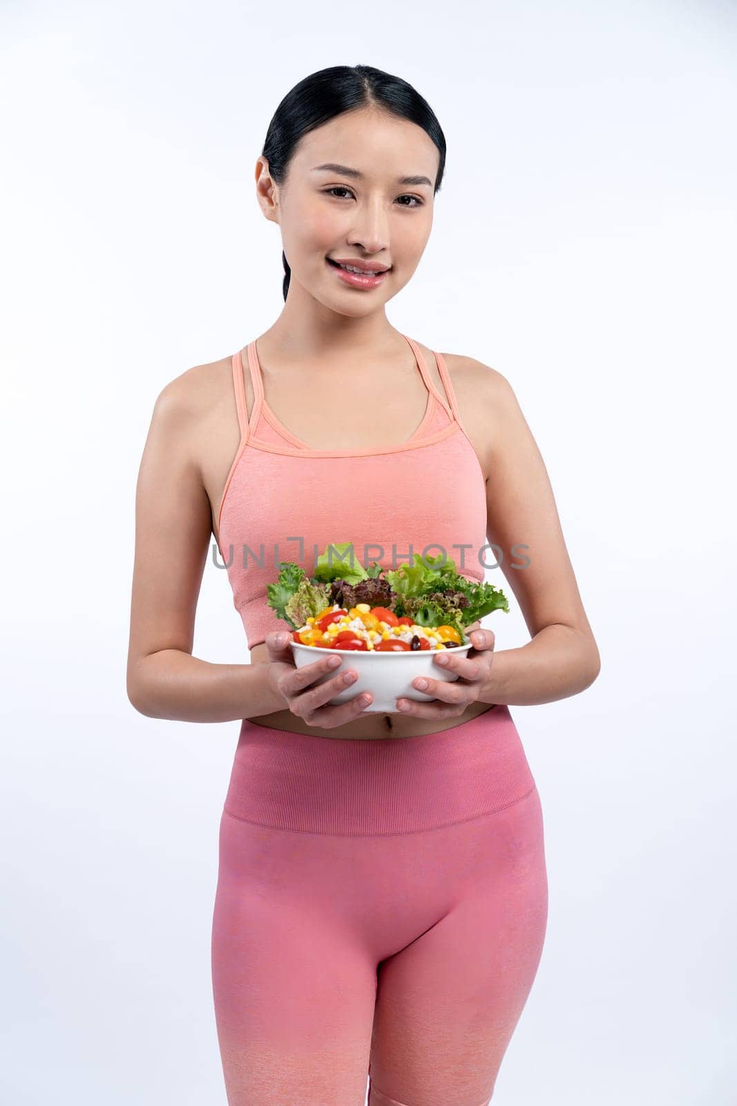 Young sporty Asian woman in sportswear holding salad bowl fill with vibrant of fruit and vegetable. Natural youthful and fit body lifestyle with balance nutrition on isolated background. Vigorous