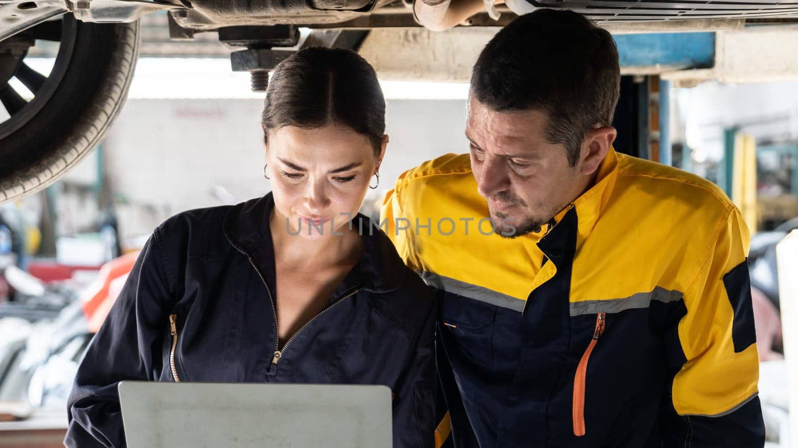 Two vehicle mechanic working together underneath lifted car, conduct car inspection with laptop. Automotive service technician in uniform carefully make diagnostic troubleshooting. Panorama Oxus