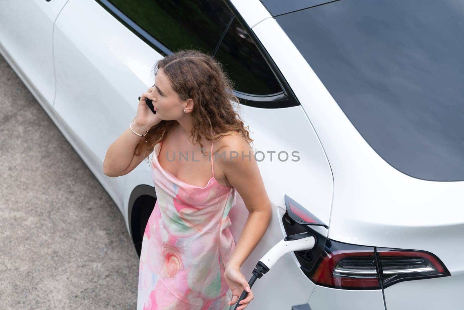 Aerial top view eco-friendly woman recharge electric vehicle from EV charging station, using EV technology utilization for tracking energy usage to optimize battery charging on smartphone. Synchronos