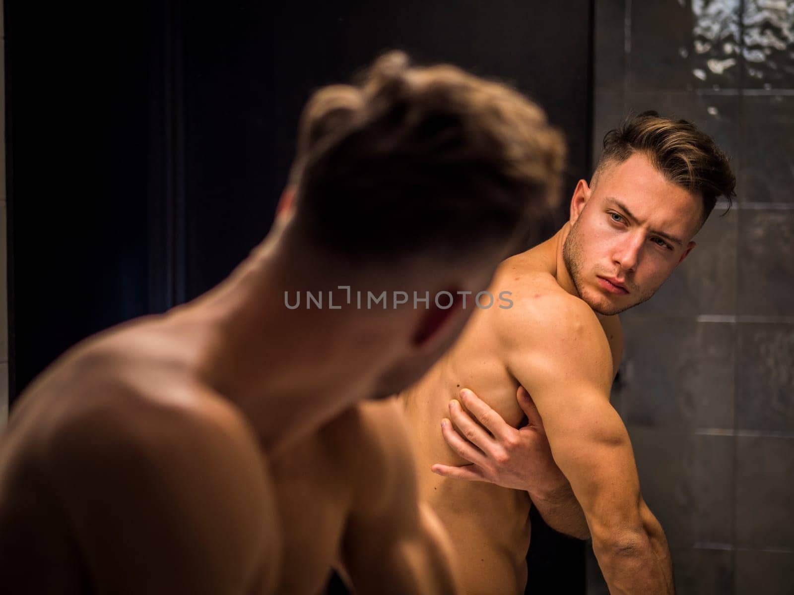 A shirtless man looking at himself in the mirror. Photo of a young man admiring his muscular physique in the mirror