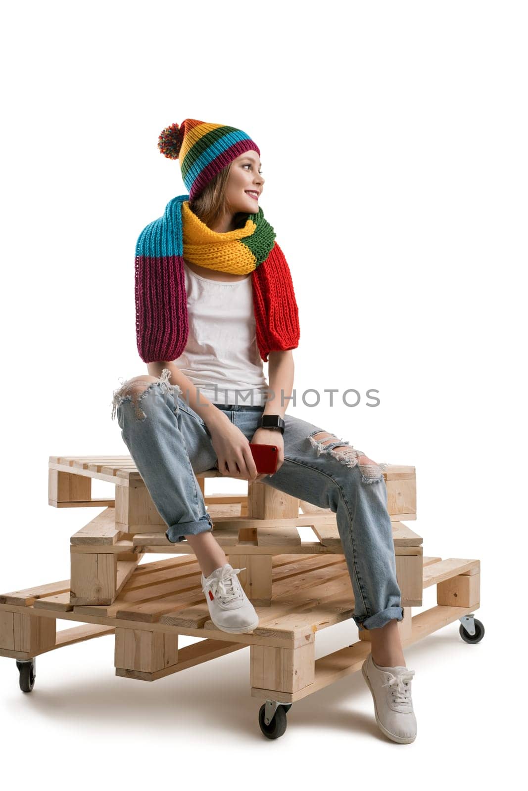 Young pretty teenager in knitted yellow hat and scarf, white t-shirt and torn jeans sitting on wooden pallets with yellow knitted bag isolated shot