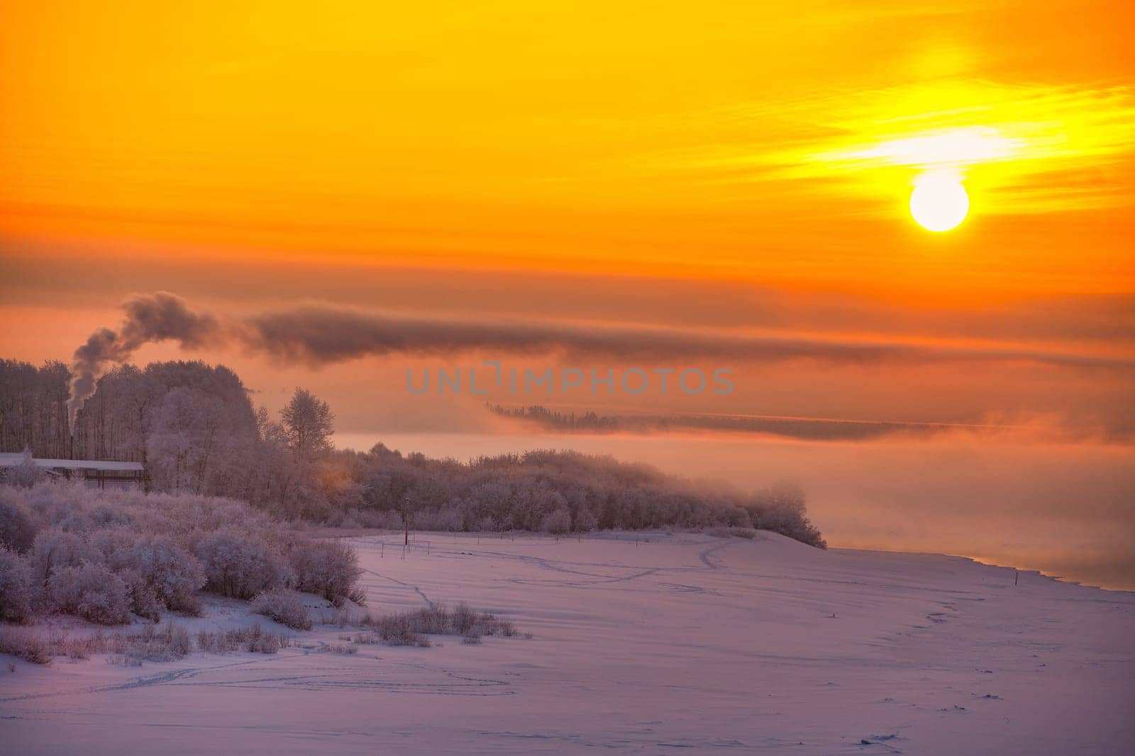 Beautiful Russian winter landscape with frozen river, bushes and houses against pink and yellow sunrise.