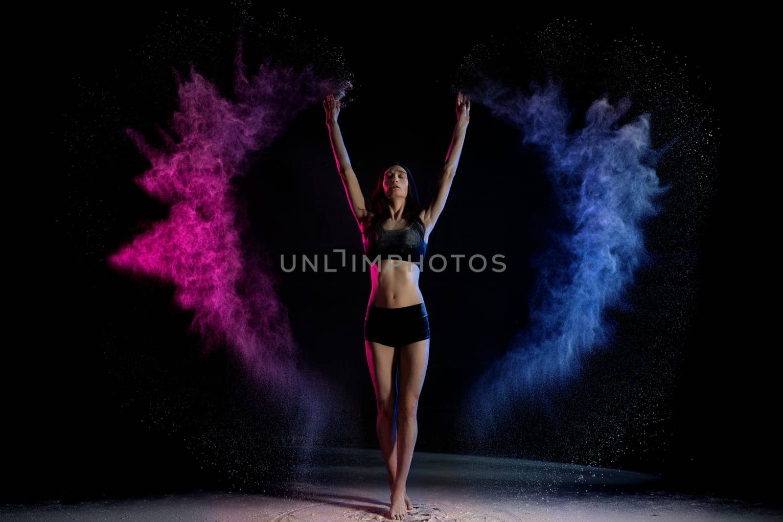 Young woman in black clothes standing with hands up in blue and pink smoke