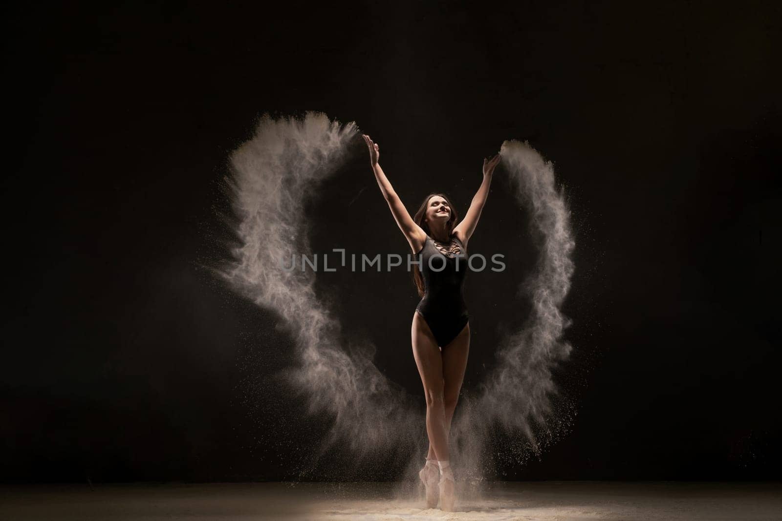 Slim ballerina in black bodysuit dancing with expression in flying white dust on white background