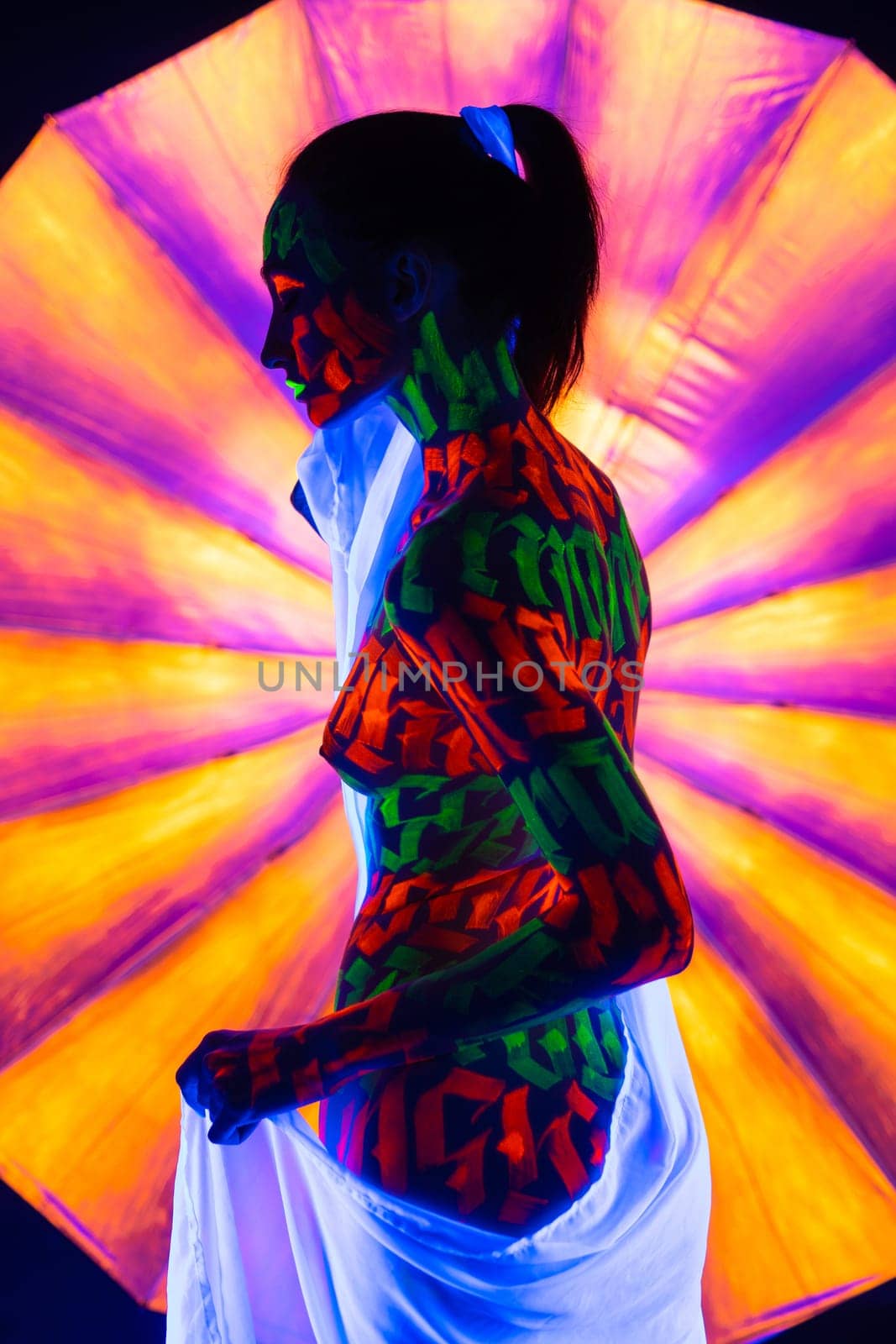 Woman with artistic UV bodyart with white ribbon in her hair and white cloth around her waist full-length reraview against colorful background