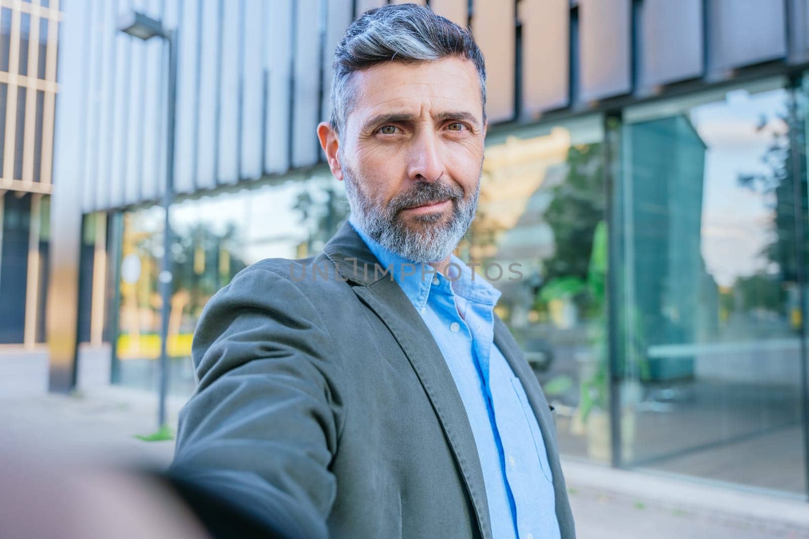 Mature businessman with gray hair and beard taking a selfie in a modern urban setting, standing near a glass office building in daylight.