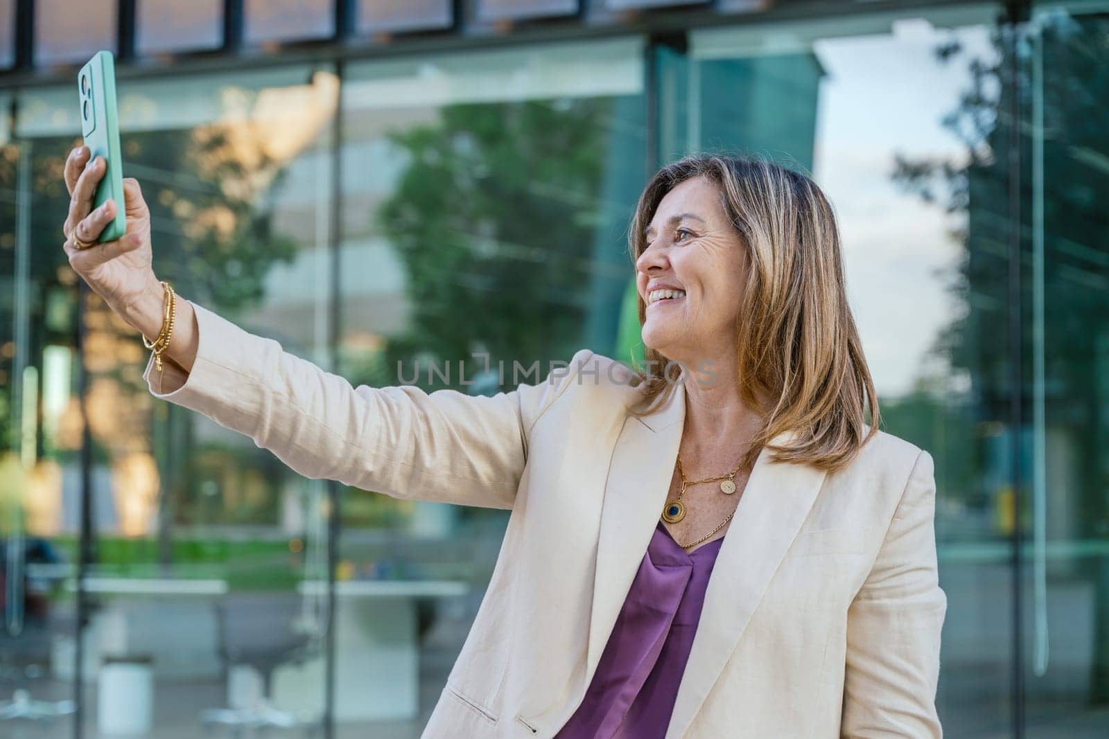 Mature businesswoman with gray hair and beard taking a selfie in a modern urban setting, standing near a glass office building in daylight.