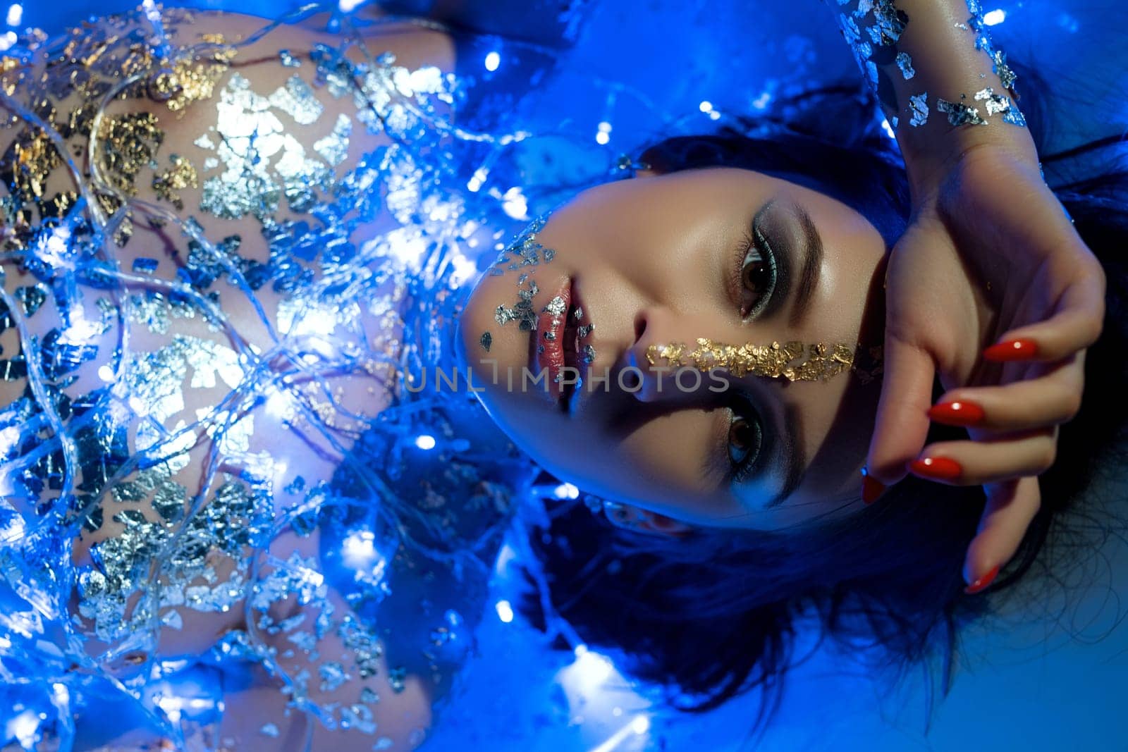 Nude girl with gold bodyart lying in the bath in blue New Year garland lights cropped close up portrait