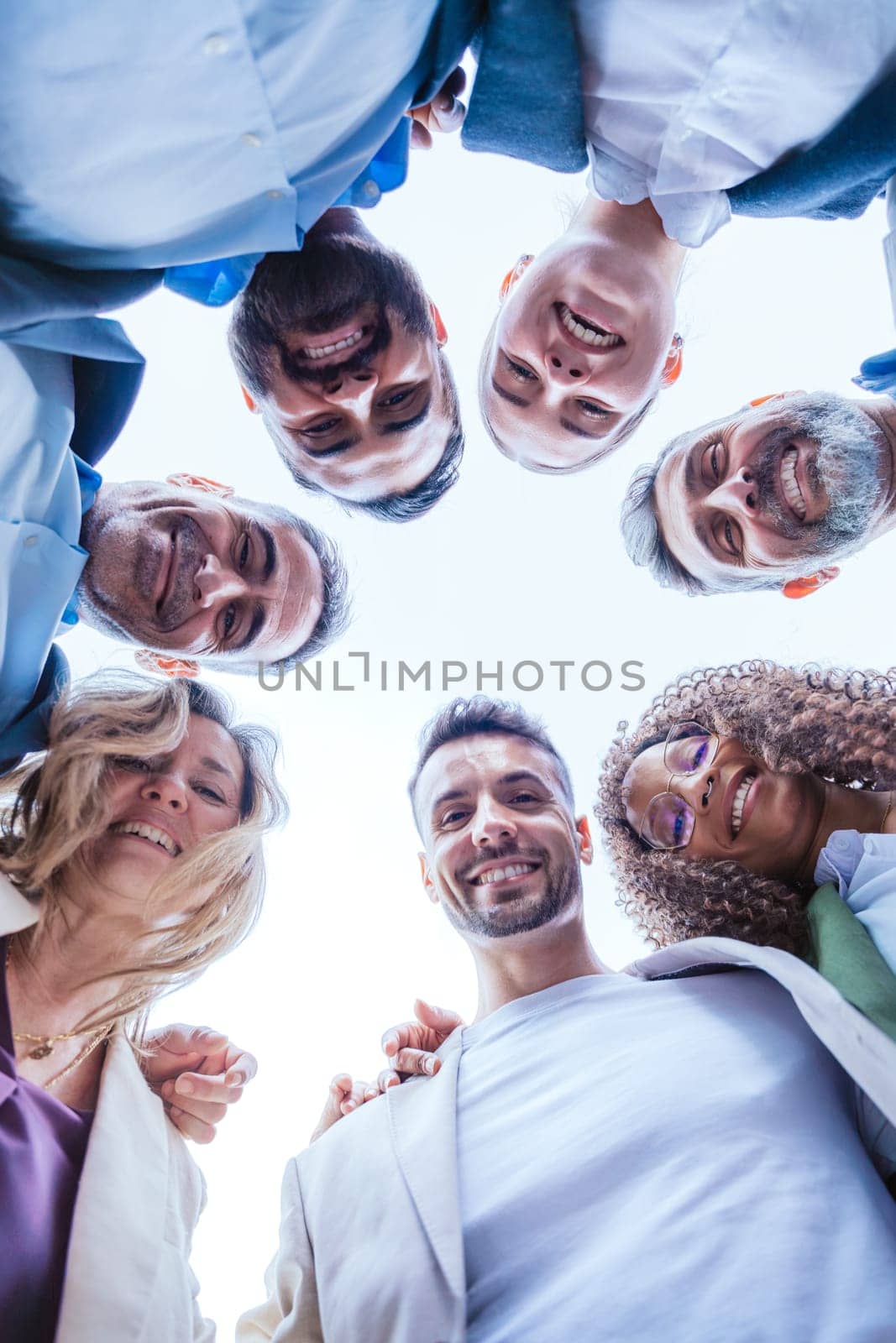 Low-angle view of a diverse group of smiling coworkers standing in a circle outdoors, symbolizing teamwork, unity, and collaboration under a clear sky.