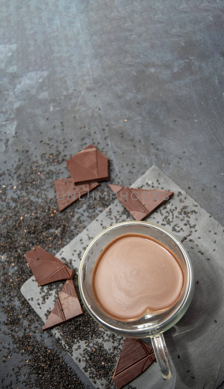 Glass cup of hot chocolate in heart shape on table surrounded by chocolate pieces and black chia seeds on grey textured background, Desert concept in color of the year 2025, mocha mousse, copy space