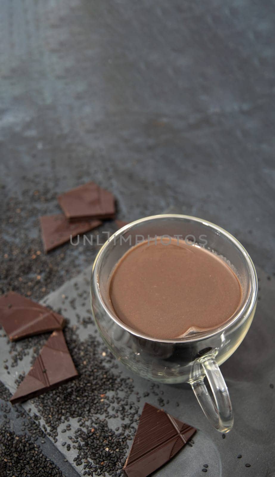 Glass cup of hot chocolate in heart shape on table surrounded by chocolate pieces and black chia seeds on grey textured background, Desert concept in color of the year 2025, mocha mousse, copy space
