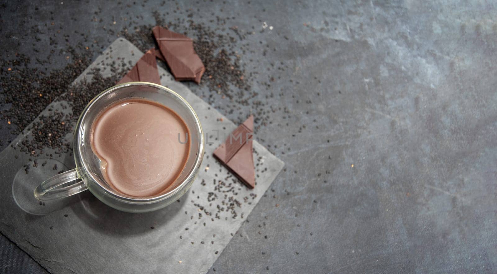 Glass cup of hot chocolate in heart shape on table surrounded by chocolate pieces and black chia seeds on grey textured background, Desert concept in color of the year 2025, mocha mousse, copy space