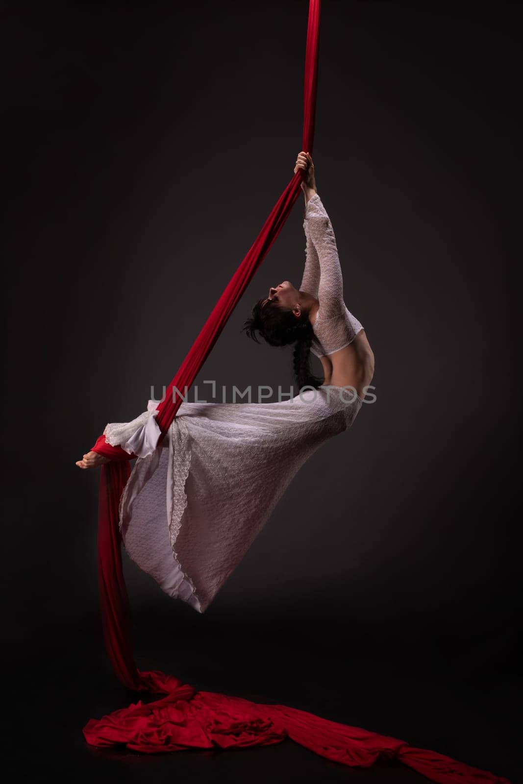 Side view of beautiful charming slim gymnast with upped hands hanging on red material on black background
