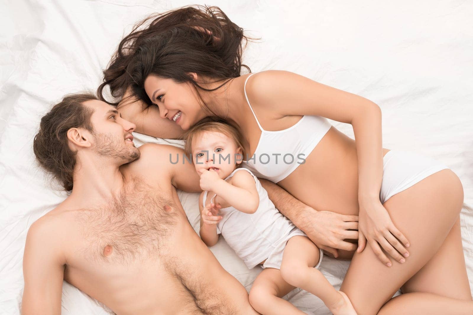 From above cheerful pregnant woman and shirtless handsome man looking at each other while lying on comfortable bed near adorable baby