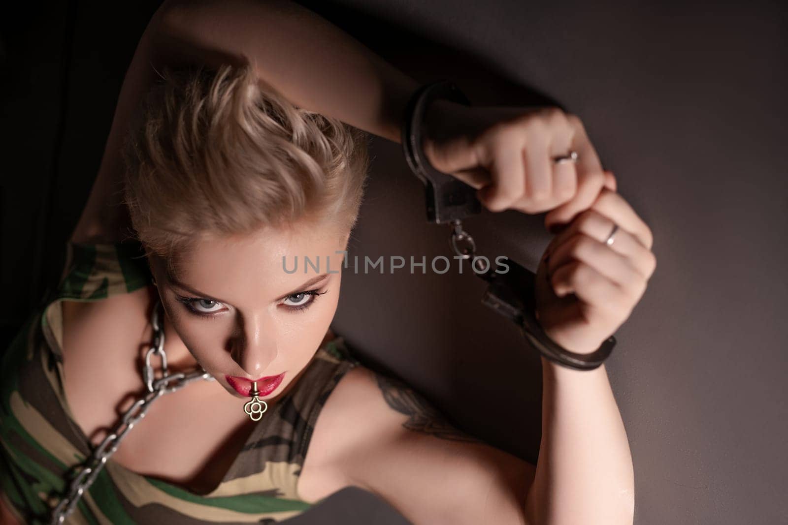 Blonde in military style top lying on the floor with a chain on her neck and wrists high angle shot in the dark room