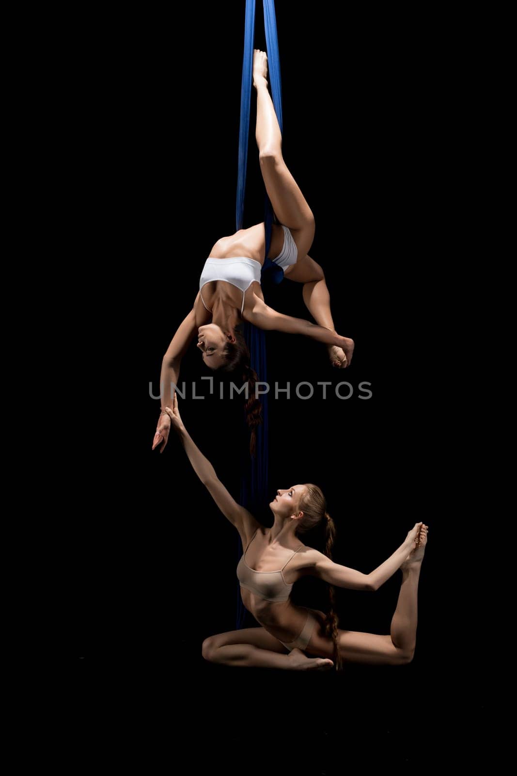 Slim woman in underwear bending body and touching hand of sensual female hanging on cloth straps while dancing against black background