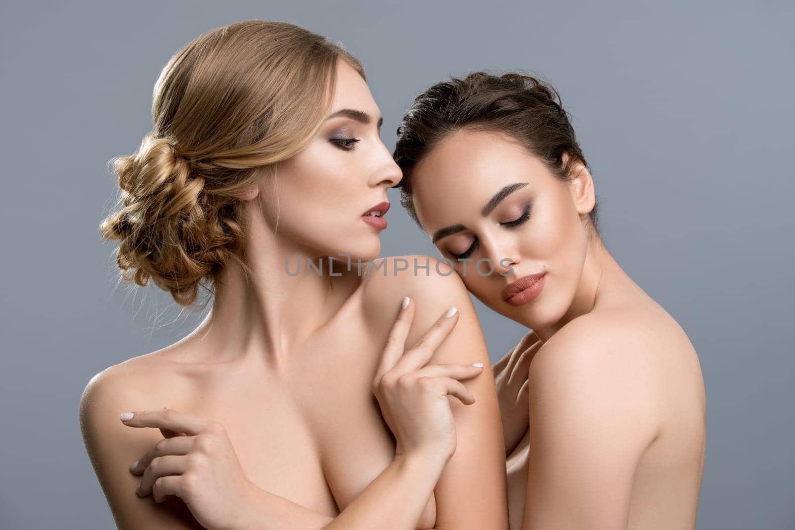 Two nude females keeping eyes closed and gently touching each other against gray background