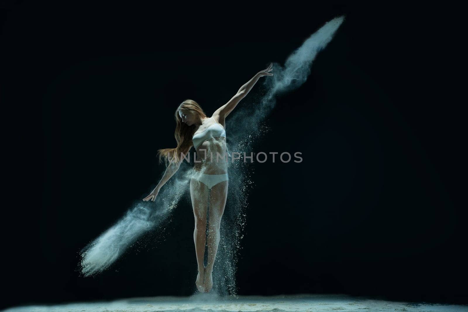Slim blonde wearing lingerie jumping gracefully in white dust cloud view in the dark room
