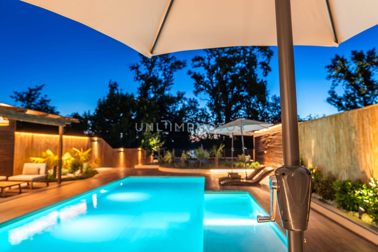 Elegant backyard pool view framed by an umbrella and serene evening sky. Modern outdoor retreat with warm lighting and stylish wooden design