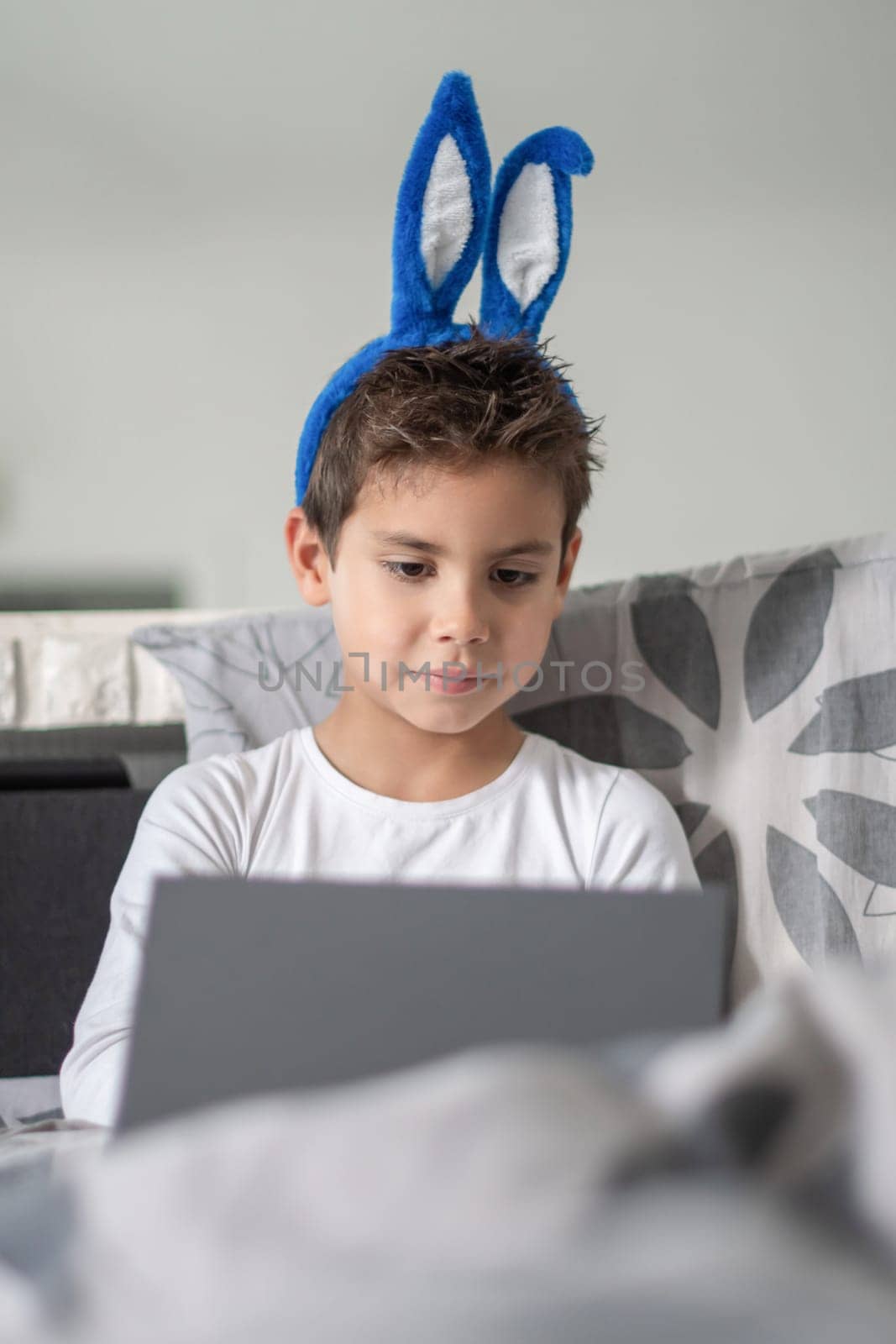 Boy with blue bunny ears sitting on sofa, concentrating on working on laptop, Casual atmosphere, playful and cheerful mood before Easter,