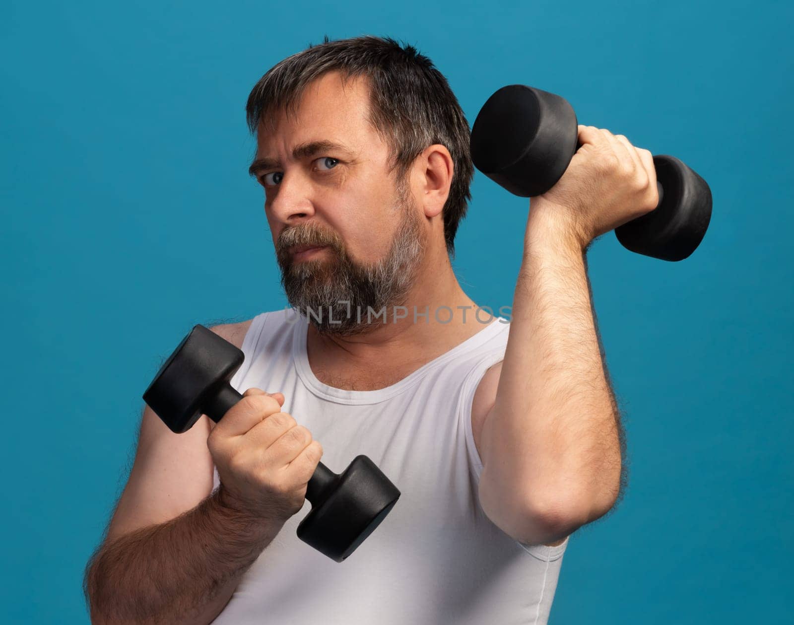 An elderly man is engaged in strength training using dumbbells, showcasing his exercise routine in a brightly colored fitness studio. The focus is on his determination and strength.